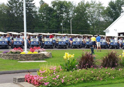 Dozens of golf carts lined up in front of a clubhouse with people preparing for a tournament, surrounded by blooming flowers and greenery.