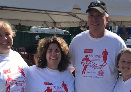 Four smiling participants wearing matching white São Paulo road race T-shirts pose together at an outdoor event under a tent.